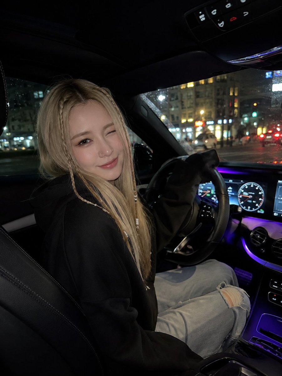 一张超写实的夜间车内自拍，年轻女子坐在驾驶座上，微微转向镜头，调皮地眨眼并带着温柔的微笑。她有一头长直的金发，脸颊两侧各编着精致的细辫，辫子上…, 图片 1