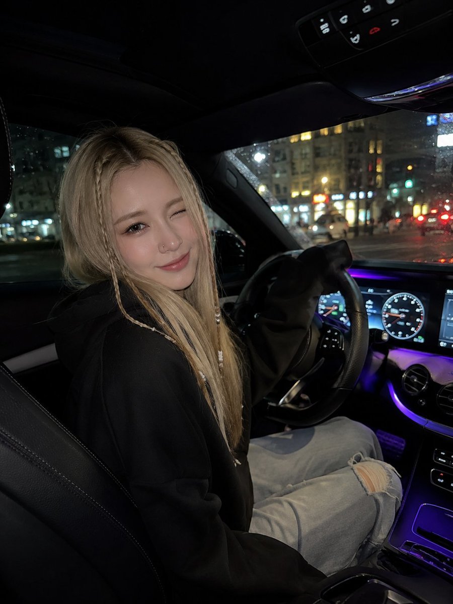 一张照片写实风格的夜间车内自拍照:一位年轻女子坐在驾驶座上,身体微微转向镜头,俏皮地眨着一只眼,露出柔和的微笑。她有一头金色长直发,脸颊两侧各…, 图片 1