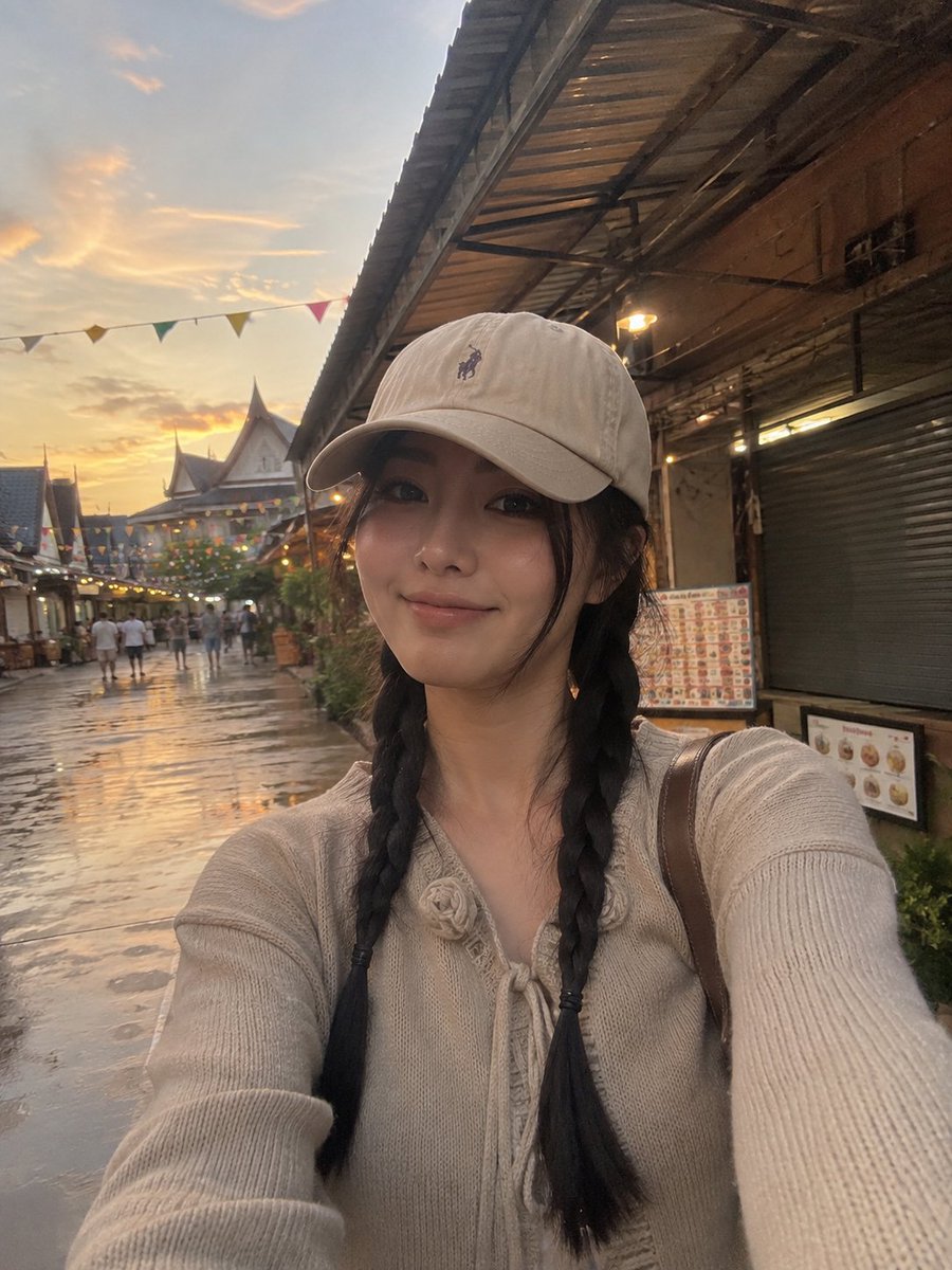 一张照片级写实的竖版旅行自拍照:一位年轻女性在黄金时段身处热闹的集市或旅游村落户外。她戴着米色棒球帽,帽子带有低调的刺绣标志;乌黑长发编成两条…, 图片 1
