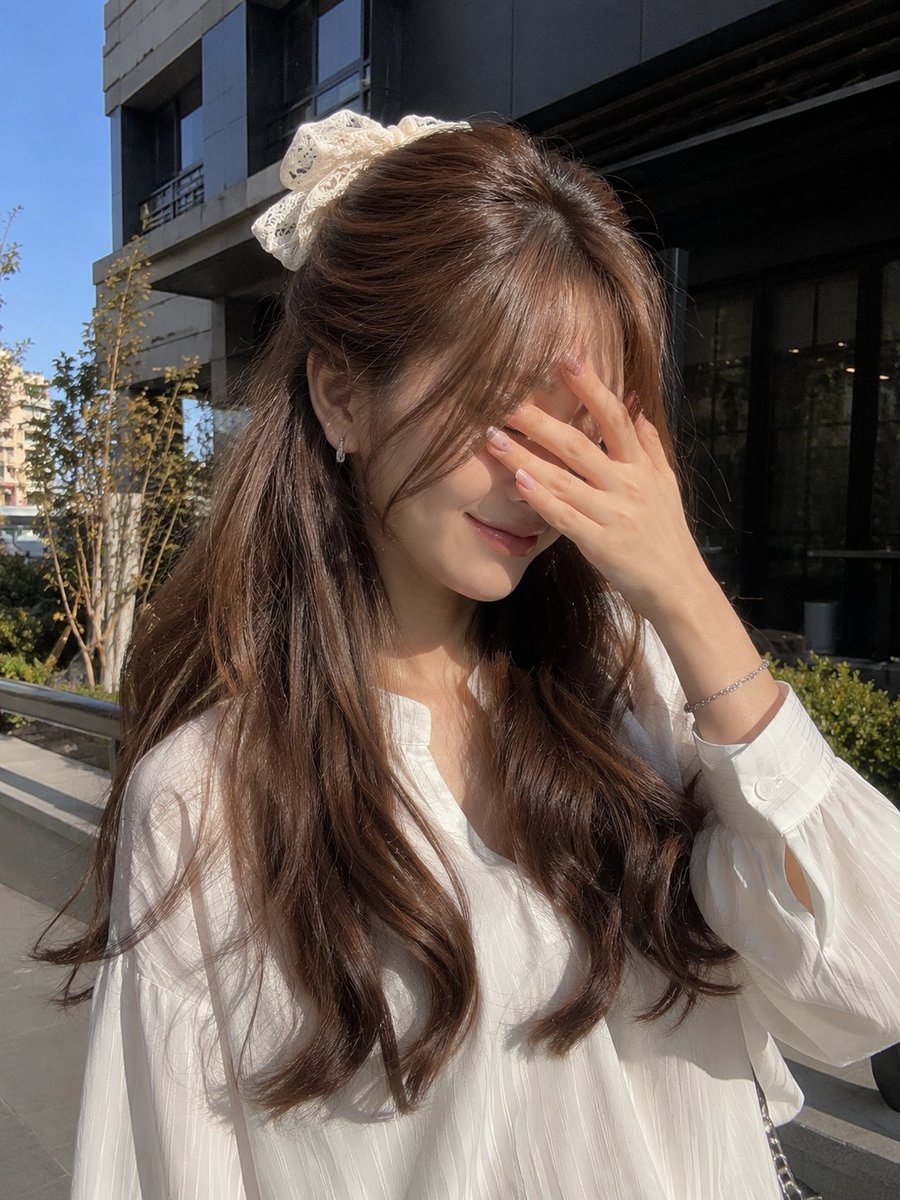 一张年轻女性的户外写实主义肖像照,她站在现代建筑外的明亮自然阳光下。她有一头浓密蓬松的浅棕色长发,呈现出柔和流畅的波浪卷,发帘式刘海轻盈地修饰…, 图片 1