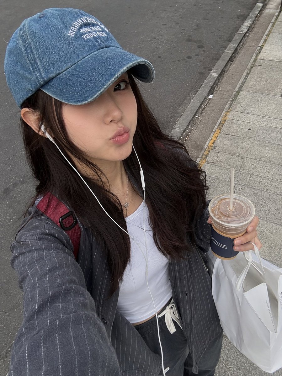 一张照片般逼真的街头风格抓拍自拍照,画面中一位年轻女性正在城市人行道上户外行走。她有一头深棕色长发,自然地垂落在褪色蓝色牛仔棒球帽下,帽子带有…, 图片 1