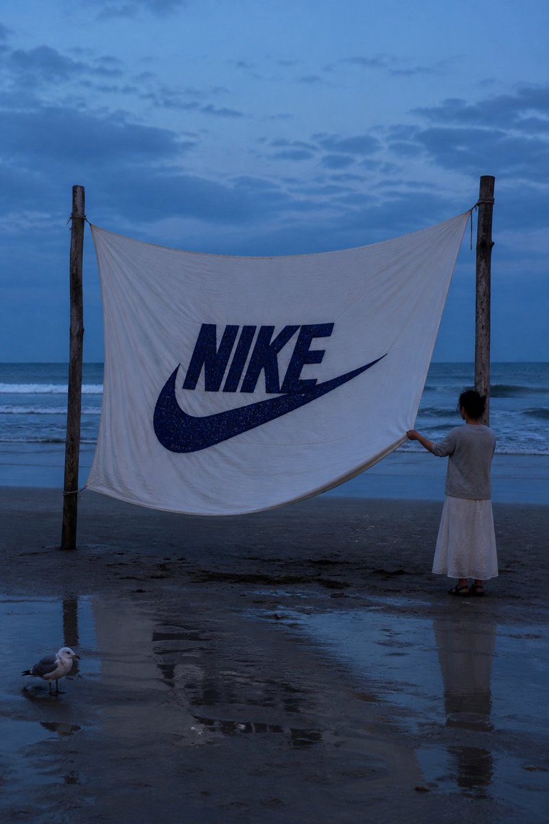 一张写实风格的海岸摄影作品。在蓝调时刻的海滩上,两根浮木杆之间悬挂着一块巨大的白布,中央用粗实的深蓝色线绣有 "NIKE" 字样及其标志,布料…, 图片 1