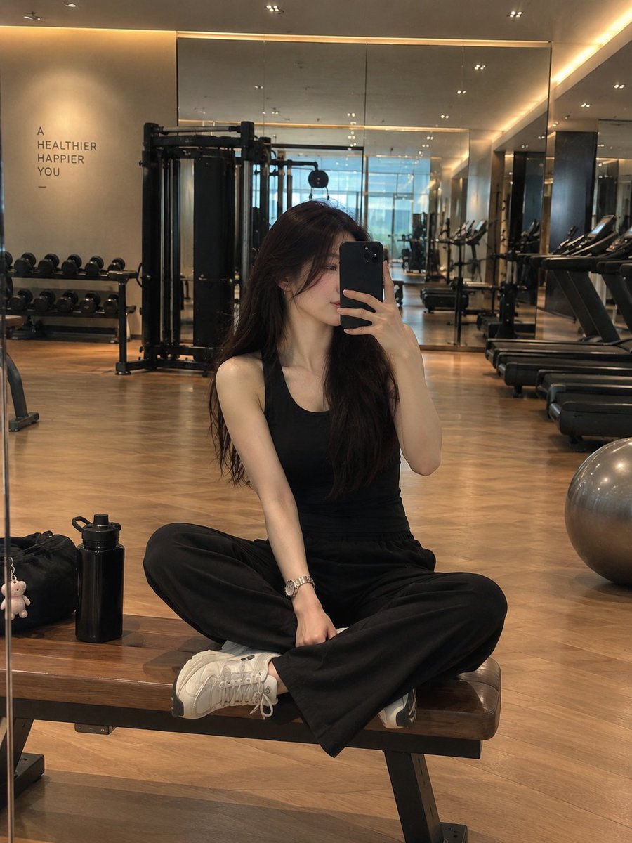 一位时尚年轻女性在现代豪华健身房内对着镜子自拍,盘腿坐在一张木质健身凳上。她留着长而顺直的深棕色头发,神情平静自然,手持一部黑色智能手机置于面…, 图片 1
