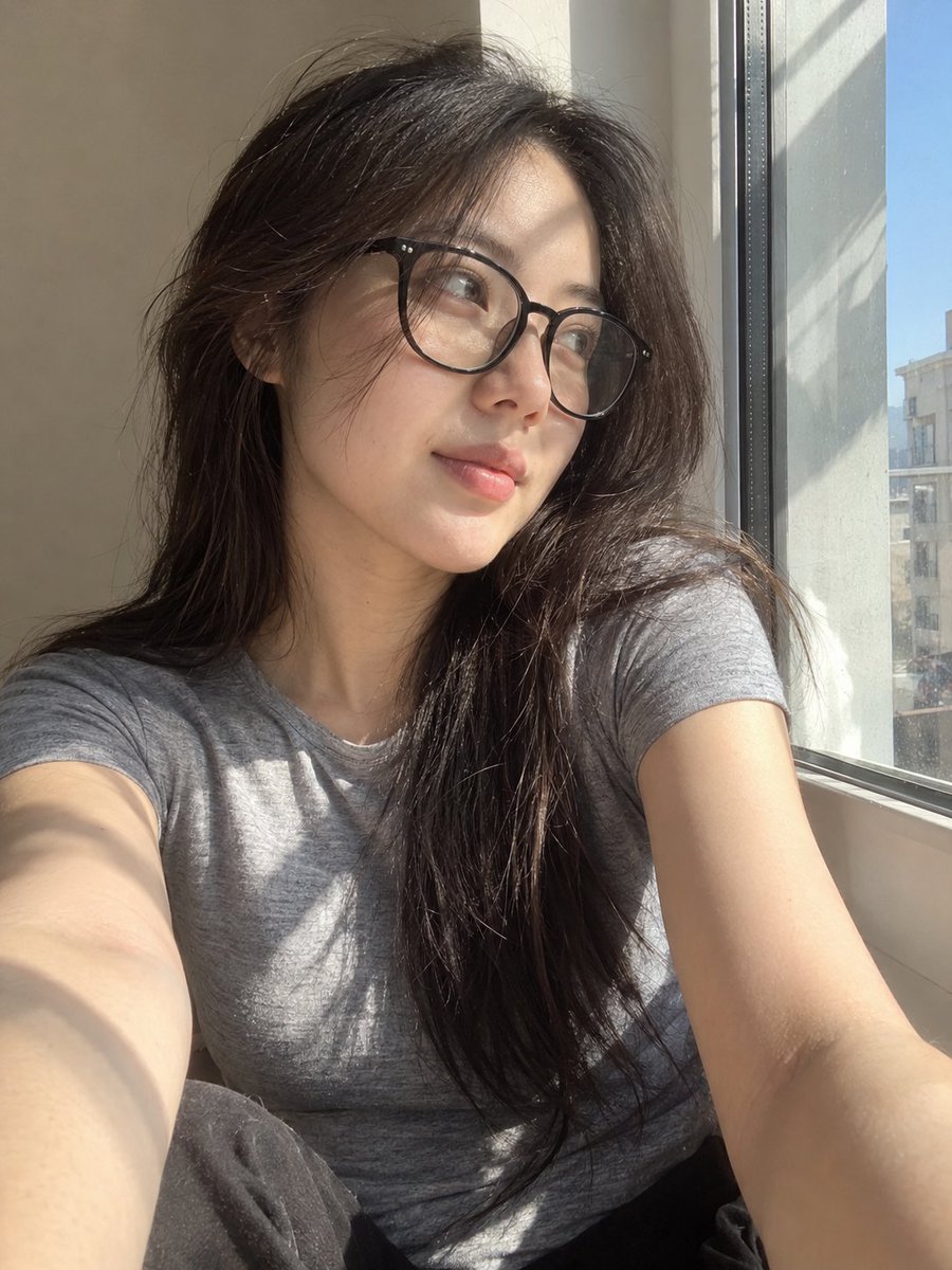 超写实风格的休闲智能手机自拍照：一位年轻东亚女性坐在安静房间内靠近阳光照射的窗户旁。保持与面部参考的高度相似性，包括柔和轮廓的鹅蛋脸、温和的下…, 图片 1