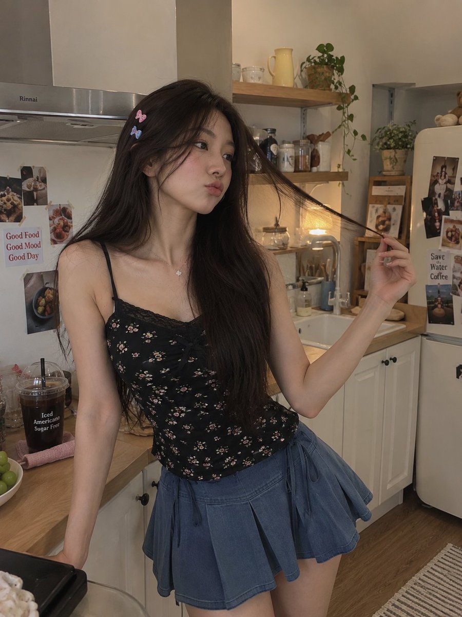 一张高质量的抓拍风格生活人像照,一位年轻女性站在温馨的现代厨房中,身穿黑色碎花细肩带吊带背心和蓝色百褶裙。她有着长而笔直的深棕色头发,别着几枚…, 图片 1