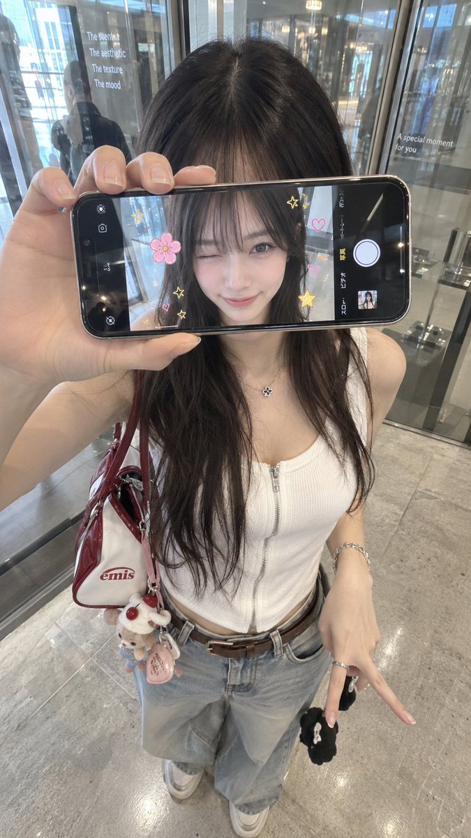 超写实 iPhone 创意人像，身份特征与参考图像完全一致。年轻女性站在室内玻璃店面或反光墙前，从略高的正面角度拍摄。她将一部智能手机水平举在…, 图片 1