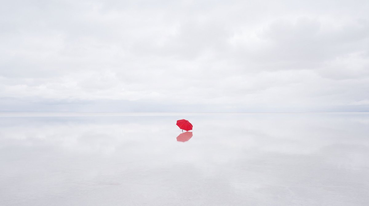 A minimal photograph of a single red umbrella on an endless pale salt flat unde…