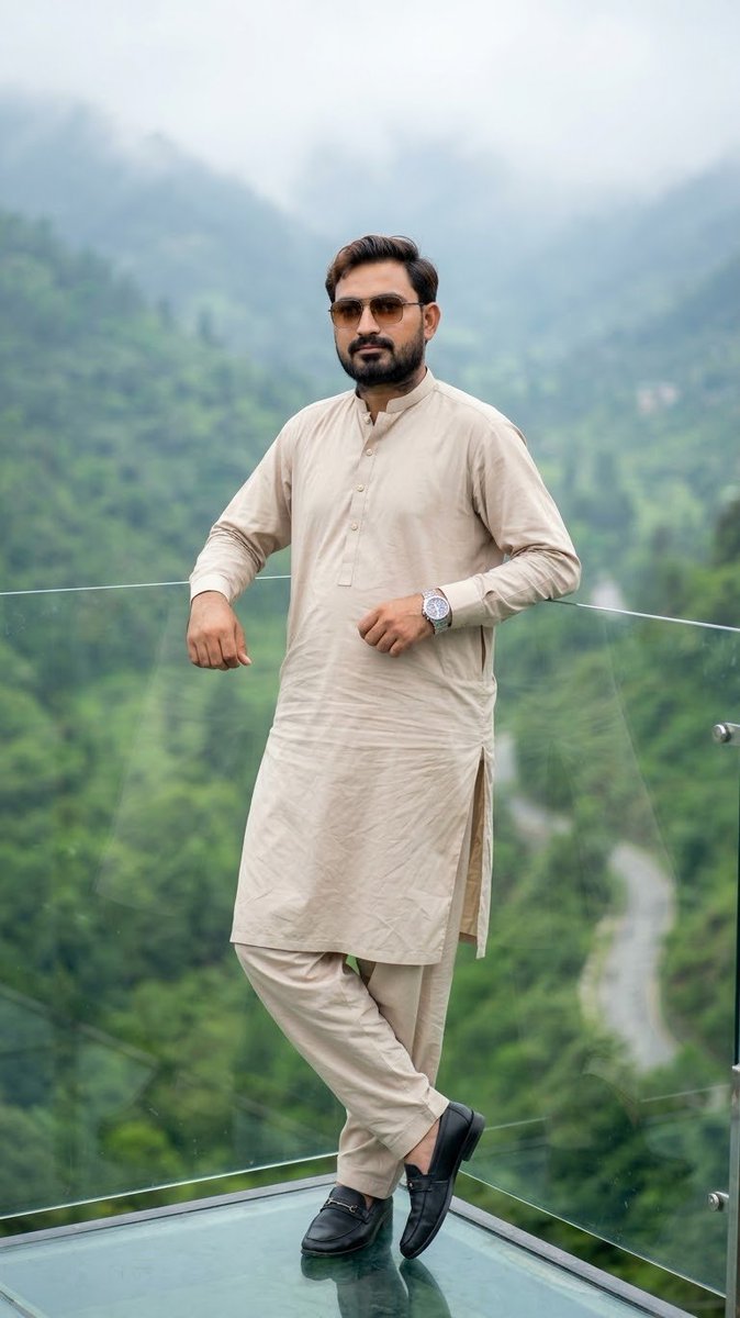 A stylish young man standing on a modern glass balcony overlooking a lush green…