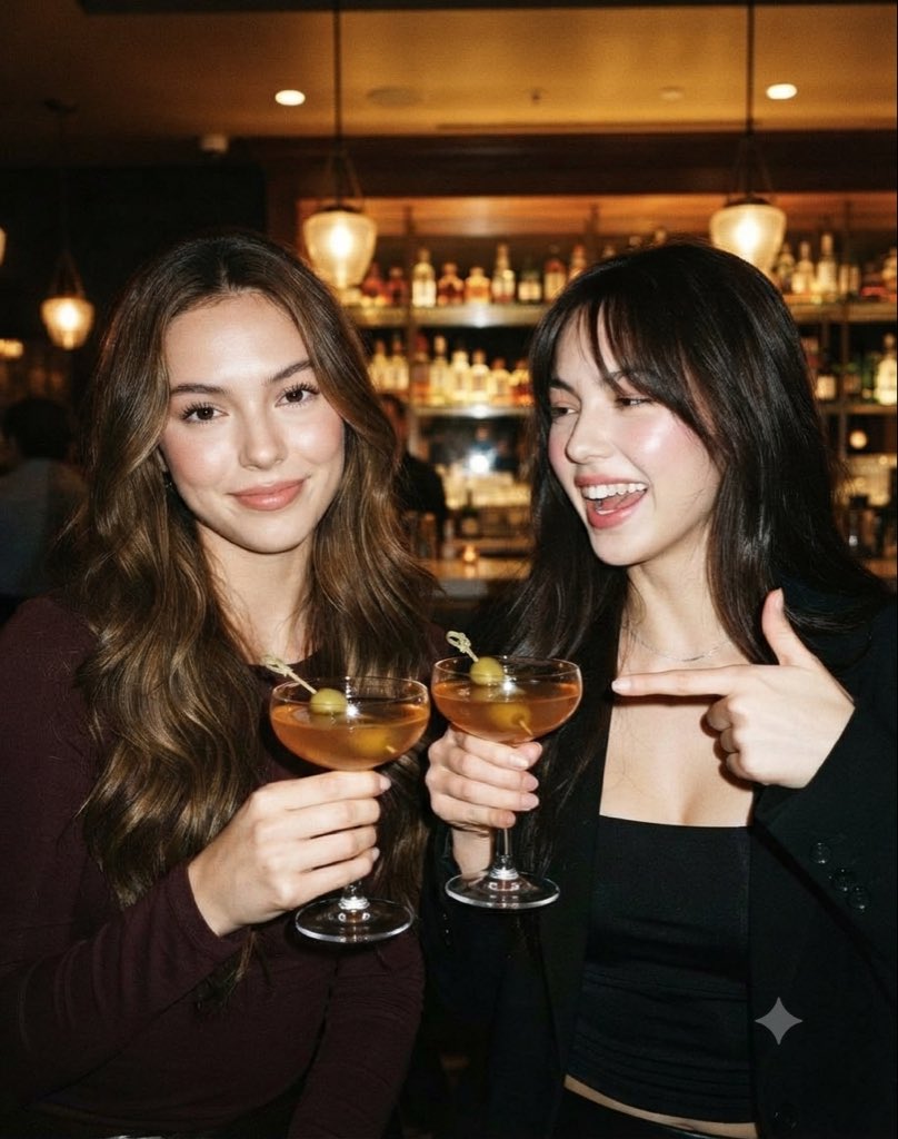 A candid, high-detail smartphone snapshot of two young women enjoying cocktails…